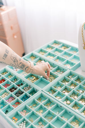 Tattooed hand reaching into mint-green compartment trays filled with gold charms, enamel pendants and colorful beads on a bright jewelry-making workstation