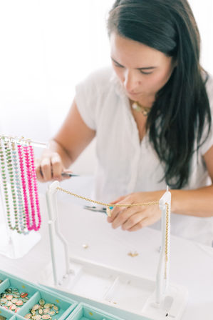 Woman artisan crafting a handmade gold chain necklace at a bright worktable, colorful beaded strands hanging on a rack and a mint organizer tray filled with charms.