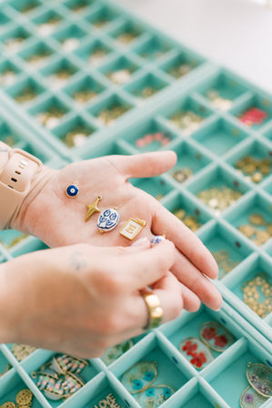 Hands holding colorful enamel and gold jewelry charms — evil eye, star, floral and letter 'E' — over a teal craft organizer tray