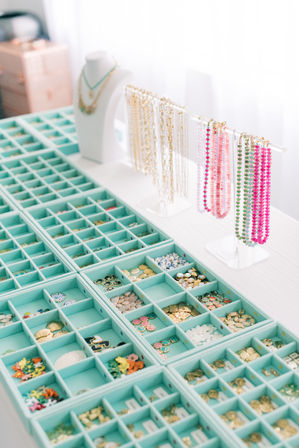 Mint-green organizer trays filled with charms, coins and beads on a white table, with gold chains and bright pink and green beaded necklaces hanging on clear display stands in a sunlit jewelry studio