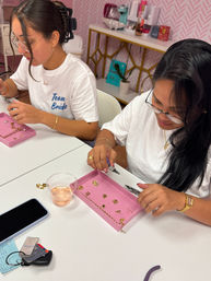 Two participants at an indoor jewelry-making workshop assembling gold charms and chains on pink trays with pliers, one wearing a 'Team Bride' tee in a pink-decor craft studio.
