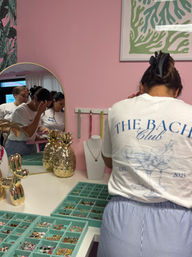 Person in a white graphic tee arranging charms at a pink jewelry boutique counter with mint-green compartment trays, gold pineapple decor, necklace bust, and a round mirror reflecting other shoppers.