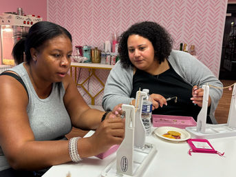 Two women at a DIY jewelry workshop in a pink studio, assembling gold chain necklaces at a white table with tools, trays, snacks and a water bottle.
