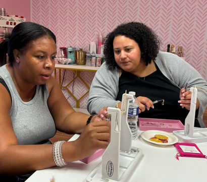 Two women at a DIY jewelry workshop in a pink studio, assembling gold chain necklaces at a white table with tools, trays, snacks and a water bottle.