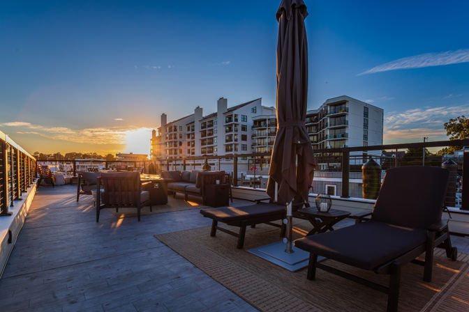 Sunset-lit waterfront rooftop lounge with cushioned sofas, chaise lounges and a closed umbrella overlooking a marina and modern condominium buildings
