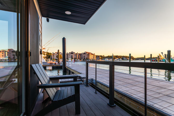Sunset view from a waterfront balcony with Adirondack chairs overlooking a marina dock, boats and waterfront condos across a calm harbor.