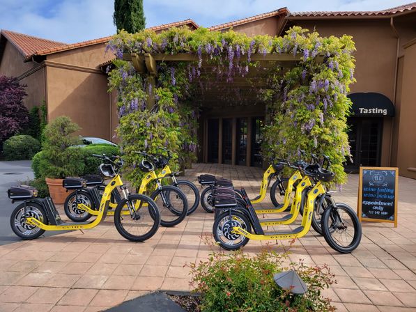 Cheerful yellow electric scooters lined up outside a wine-tasting entrance draped in purple wisteria on a sunlit terracotta-tiled plaza.
