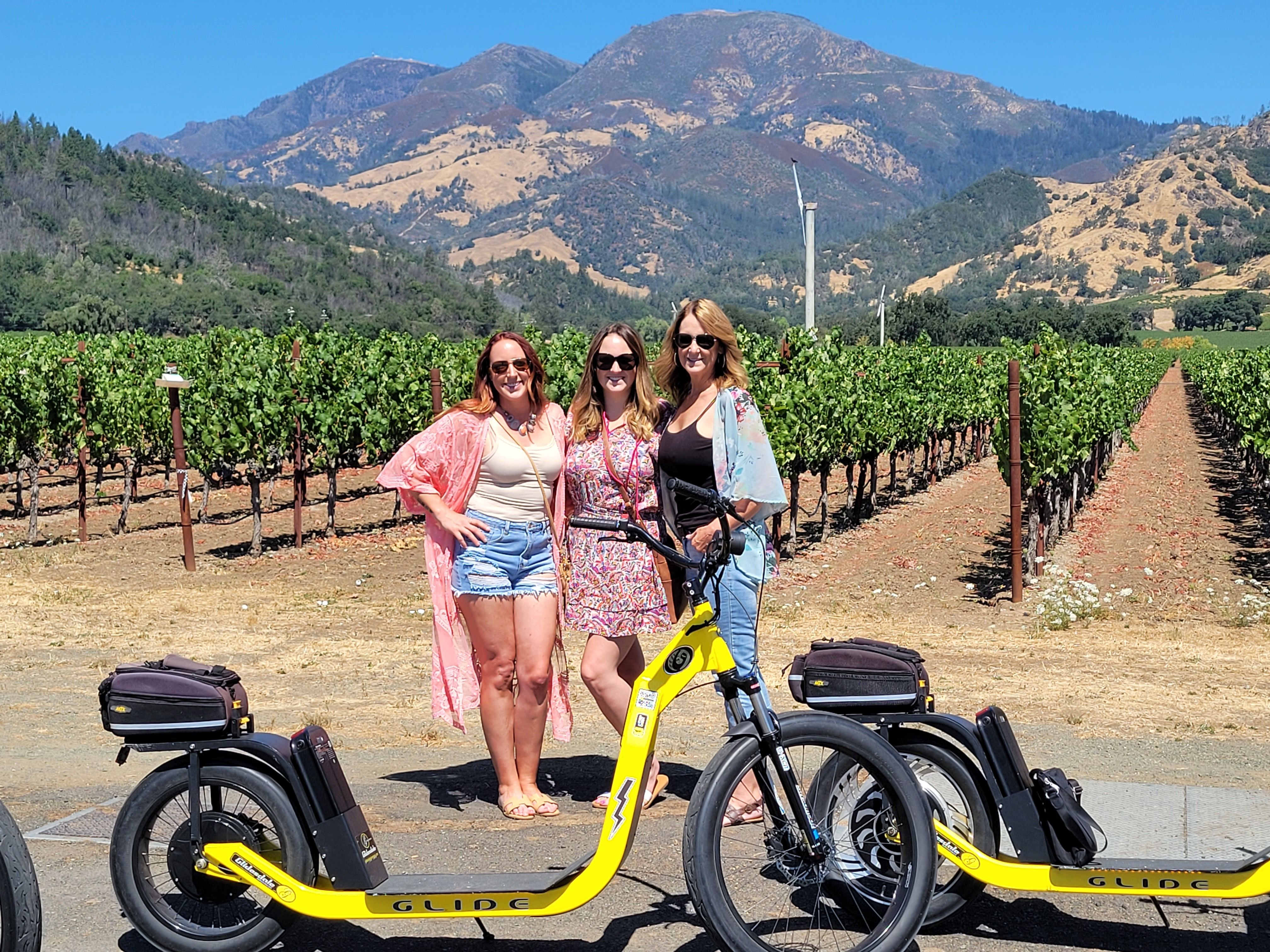 Three women in summer outfits and sunglasses pose with yellow electric scooters in a sunny vineyard, rows of grapevines leading to golden hills and blue mountains under a clear sky.