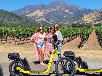 Three women in summer outfits and sunglasses pose with yellow electric scooters in a sunny vineyard, rows of grapevines leading to golden hills and blue mountains under a clear sky.