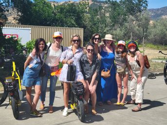 Eight women in casual summer outfits smiling and posing with yellow electric bikes on a sunny hillside bike tour, trees and dry hills in the background.