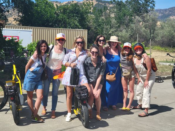 Eight women in casual summer outfits smiling and posing with yellow electric bikes on a sunny hillside bike tour, trees and dry hills in the background.
