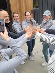 Small group toasting with rosé champagne flutes on an outdoor winery tasting patio, smiling and clinking glasses in casual jackets and sneakers.