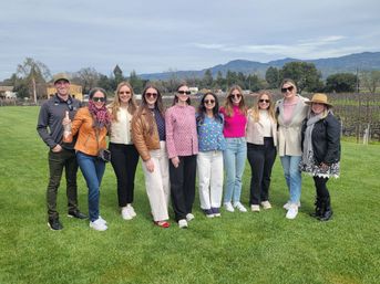 Eleven friends posing on a green lawn at a vineyard in wine country with grapevines and mountains behind them, casual spring outfits and sunglasses, one holding a bottle.