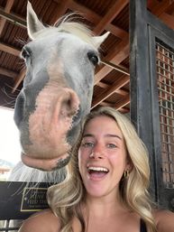 Smiling woman taking a close-up selfie with a curious gray horse poking its nose through a wooden barn stall