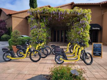 Bright yellow electric scooters lined up outside a wisteria-covered winery tasting room with terracotta roofs and a chalkboard sign