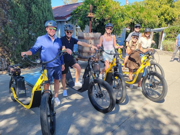 Six adults in helmets posing on yellow electric scooters during a sunny wine-tour stop in a winery courtyard with a wooden wine press