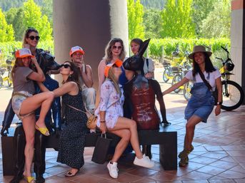 Group of seven women in summer outfits and orange caps posing and laughing with bronze rabbit sculptures on a bench under a shaded tiled patio, with electric bikes and a sunlit vineyard and trees in the background.