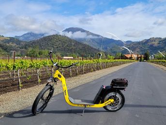 Yellow electric scooter parked on a paved road beside lush vineyard rows, with rolling green mountains and low clouds under a blue sky — scenic wine-country landscape.