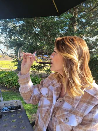 Woman sipping red wine from a large glass at an outdoor garden patio table under an umbrella, surrounded by evergreen trees and lawn with a smartphone on the table — relaxed afternoon vibe