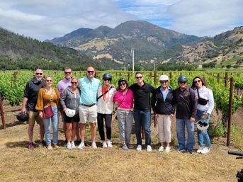 Smiling group of people in casual clothes and helmets posing in a sunlit vineyard with neat rows of grapevines and rolling mountains under a partly cloudy sky — outdoor wine-country photo.