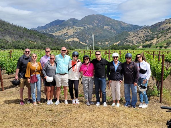 Smiling group of people in casual clothes and helmets posing in a sunlit vineyard with neat rows of grapevines and rolling mountains under a partly cloudy sky — outdoor wine-country photo.