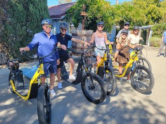 Six adults in helmets smiling with bright yellow electric scooters on a sunny wine-country courtyard beside a wooden wine press and leafy trees.