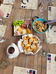 Top-down view of a rustic wooden wine-tasting table with a charcuterie board (green grapes, sliced cheeses, cured meats, nuts), a blue bowl of pasta salad, a basket of baguette slices, a ceramic pitcher, wine glasses and tasting menus.