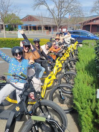 Cheerful group of riders on yellow electric bikes lined up in a sunny small‑town main street parking area near shops and bare trees, ready for an e-bike tour.