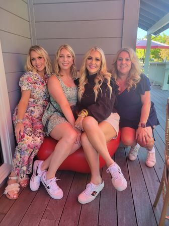 Four smiling women in summer dresses and sneakers sitting close on a red bench on a sunny wooden patio with umbrellas and greenery in the background