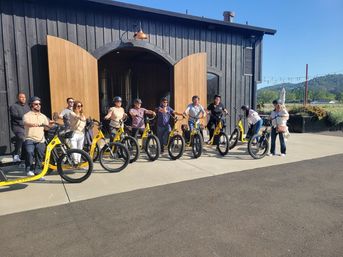 Group of people lined up on yellow electric scooters outside a rustic black barn with open wooden doors, ready for a sunny wine-country e-scooter tour with rolling hills in the background