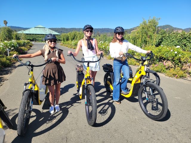 Three women in helmets smiling on yellow electric bikes, posing on a sunny paved vineyard road framed by grapevines, rolling hills and a clear blue sky — lively wine-country e-bike outing.