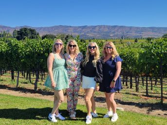 Four women in summer dresses and sunglasses posing in a sunlit vineyard with rows of grapevines and distant mountains under a clear blue sky — wine country outdoor portrait.
