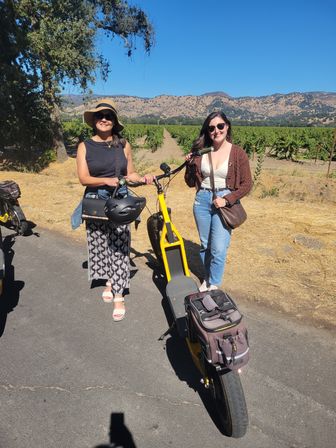Two women in sunglasses posing with a yellow electric scooter on a sunny vineyard road, grapevines stretching toward rolling hills under a clear blue sky