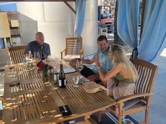 Three adults sharing wine at a sunlit outdoor patio wooden table with a bouquet centerpiece, wine bottles and glasses, and blue curtains, engaged in animated conversation