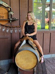 Woman in a black tank top sitting on a leather saddle mounted on a wine barrel at a rustic winery patio, wooden wall with hanging wine glasses and a bottle, window showing outdoor seating and greenery, smiling.