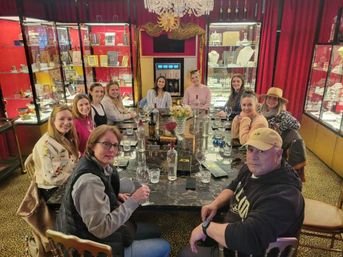 Group of a dozen people seated around a marble tasting table with wine glasses and bottles in a luxurious boutique showroom featuring red velvet curtains, glass display cabinets, a chandelier and ornate mirror.