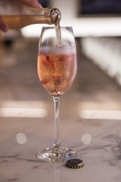 Pouring sparkling rosé into a champagne flute with pomegranate seeds — effervescent pink cocktail on a marble bar counter with bottle cap and soft bokeh background.