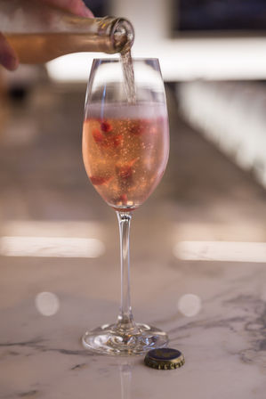 Pouring sparkling rosé into a champagne flute with pomegranate seeds — effervescent pink cocktail on a marble bar counter with bottle cap and soft bokeh background.