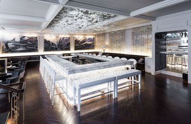 Upscale modern marble U-shaped bar glowing with backlit white stools and tufted benches, dark herringbone wood floor, abstract wall art and a sculptural glass ceiling installation in a luxury lounge