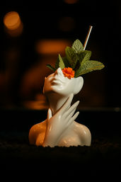 Stylized white ceramic bust cocktail mug with a metal straw, fresh mint leaves and an orange marigold garnish, hand cradling the neck against a moody dark bokeh bar background.