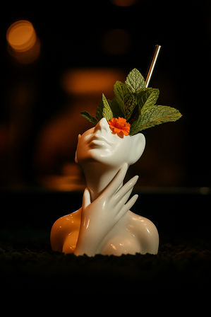 Stylized white ceramic bust cocktail mug with a metal straw, fresh mint leaves and an orange marigold garnish, hand cradling the neck against a moody dark bokeh bar background.