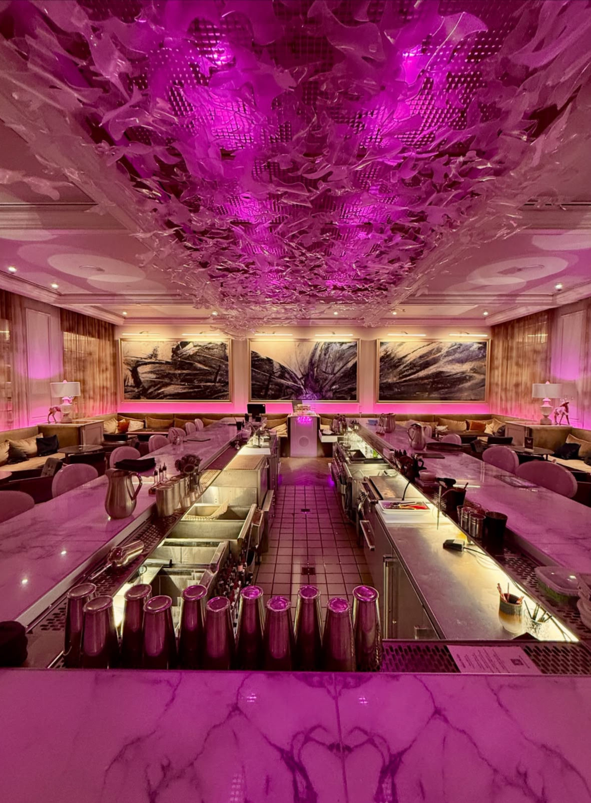 Luxury hotel bar lounge interior with long marble island, row of cocktail shakers, sculptural ceiling installation and vibrant magenta lighting over plush seating and abstract wall art