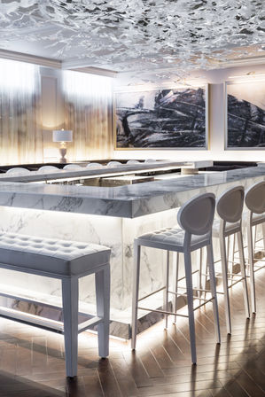 Luxury marble bar interior in a modern hotel, featuring an illuminated white marble island, tufted bench and tall oval-back bar stools, sculptural glass ceiling installation, abstract wall art and herringbone wood floor.