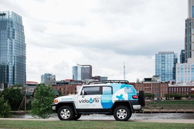 Blue-and-white branded SUV parked on a riverside lawn with a downtown glass-and-brick skyline behind under a cloudy sky.