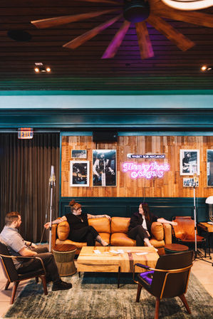 Cozy boutique hotel lounge with three people chatting on a caramel leather sofa and armchairs around a wooden coffee table, wood-paneled wall with framed photos and a pink neon sign, warm lighting and ceiling fan