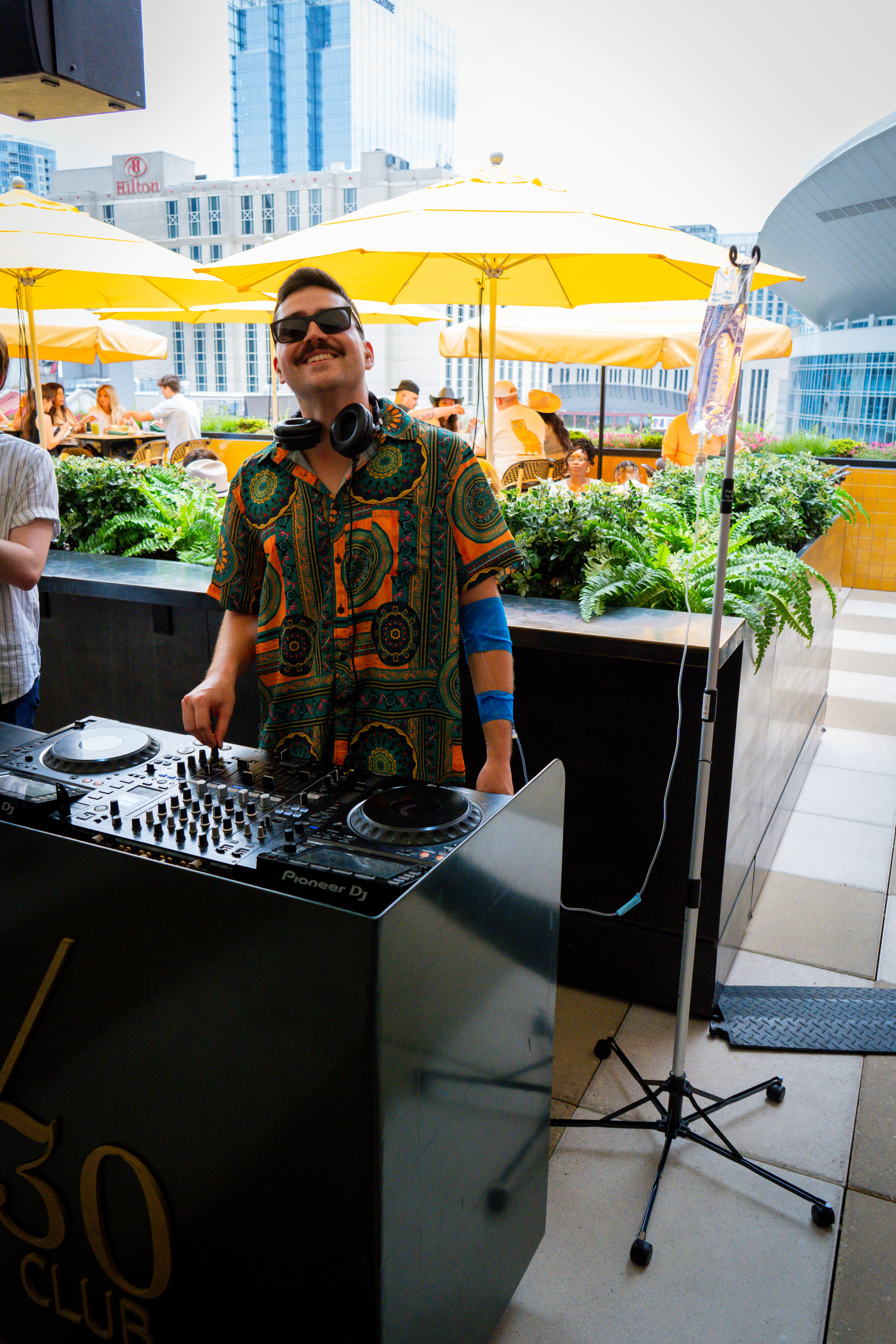 Cheerful DJ in a colorful patterned shirt and sunglasses spinning at a rooftop terrace DJ booth with turntables, bright yellow umbrellas, lush planters and urban skyscrapers in the background; a tall stand with tubing nearby.