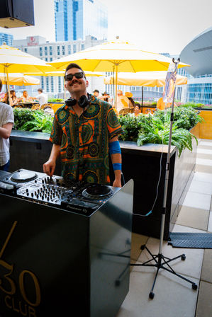 Cheerful DJ in a colorful patterned shirt and sunglasses spinning at a rooftop terrace DJ booth with turntables, bright yellow umbrellas, lush planters and urban skyscrapers in the background; a tall stand with tubing nearby.