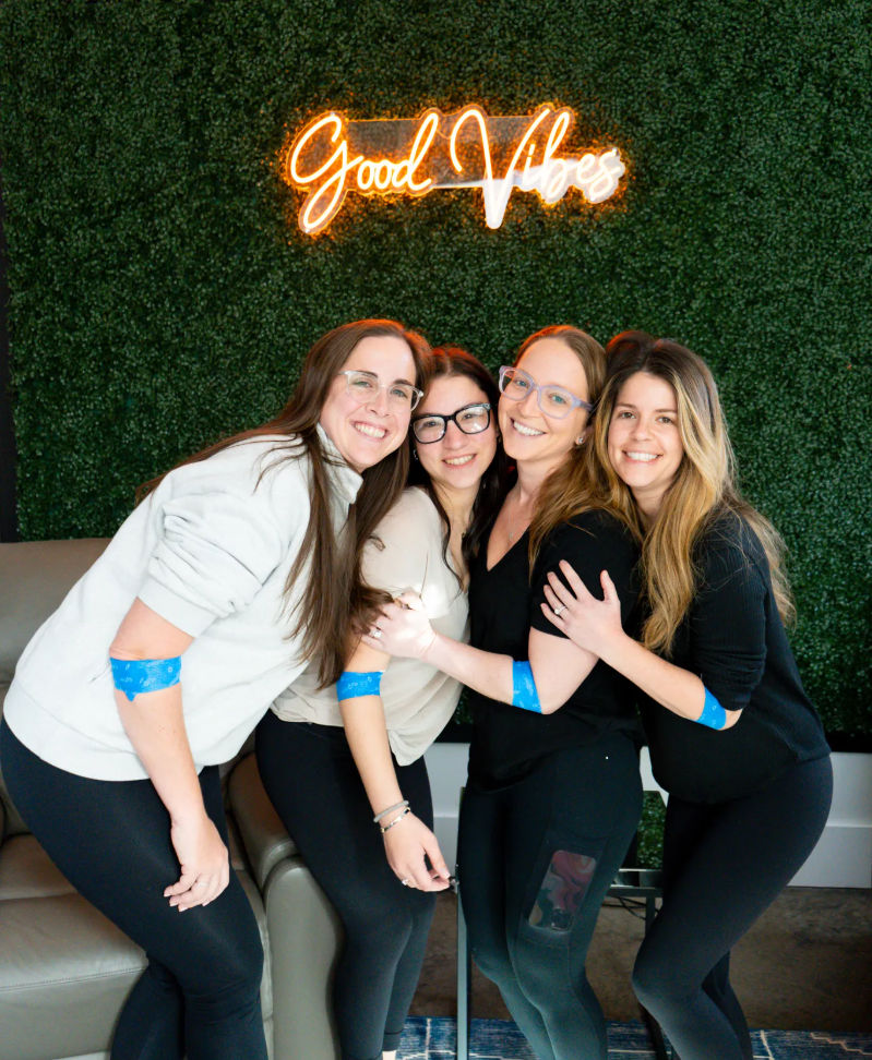 Four smiling women in casual activewear hugging indoors in front of a green hedge wall with a neon “Good Vibes” sign, each wearing bright blue adhesive arm bandages.