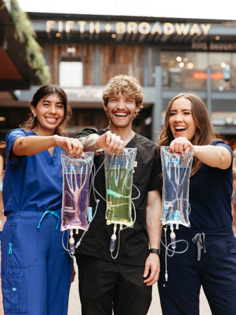 Three smiling healthcare workers in scrubs hold colorful IV hydration bags (pink, green, clear) in an outdoor urban plaza, showcasing IV therapy, wellness, and medical staff camaraderie.