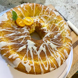 Glazed lemon bundt cake with white icing drizzle, kumquat halves and mint garnish, one slice removed, on a white plate atop a wooden cutting board